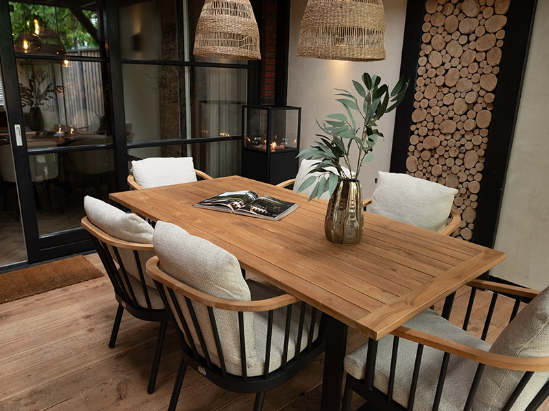 Sfeervolle overdekte veranda met houten eettafel, comfortabele stoelen met kussens, rotan hanglampen en decoratieve houtstapelwand, ontworpen door Nouk Interiors.