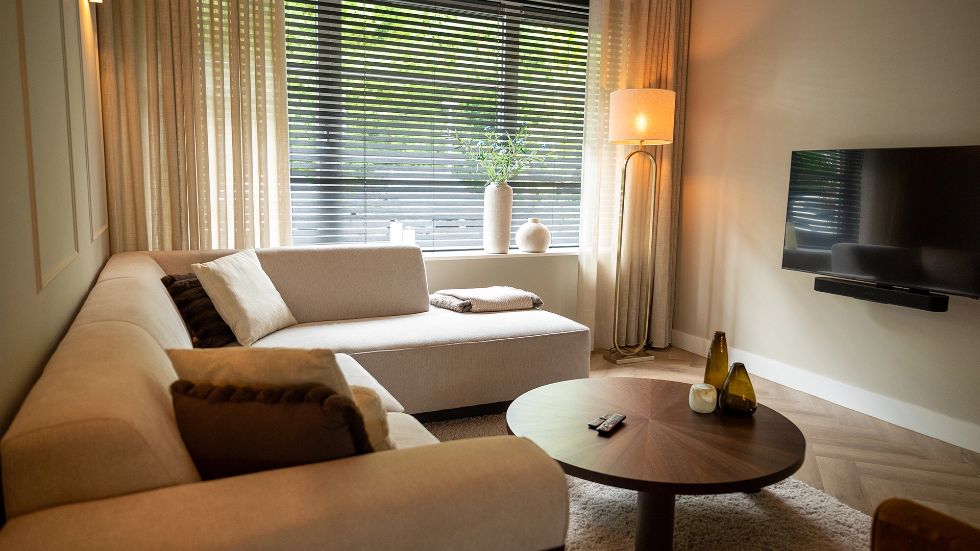 Gezellige woonkamer met lichte hoekbank, houten salontafel, grote raampartij met linnen gordijnen en zwarte jaloezieën, ontworpen door Nouk Interiors.