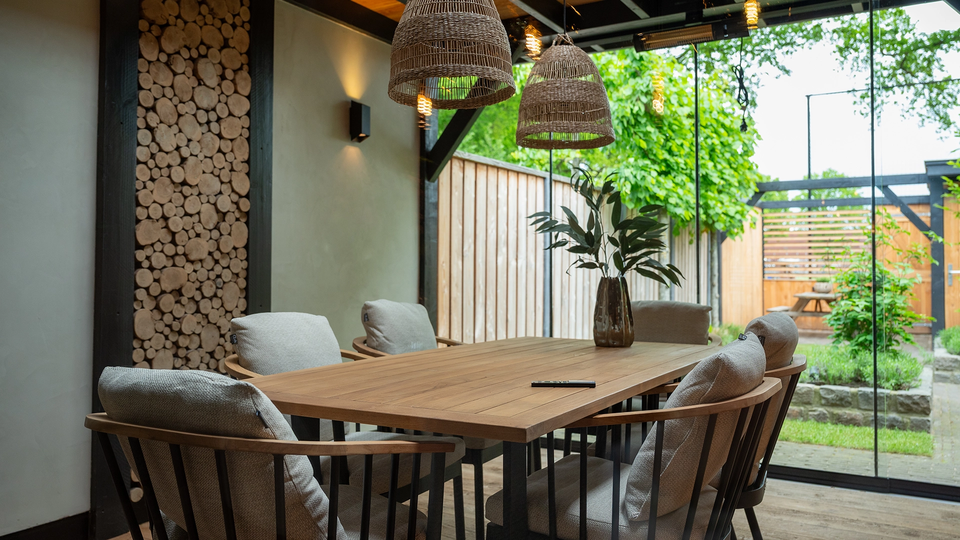 Overdekte veranda met houten eettafel, comfortabele stoelen en rotan hanglampen, gecombineerd met glazen schuifwanden en tuinontwerp, gerealiseerd door Nouk Interiors.