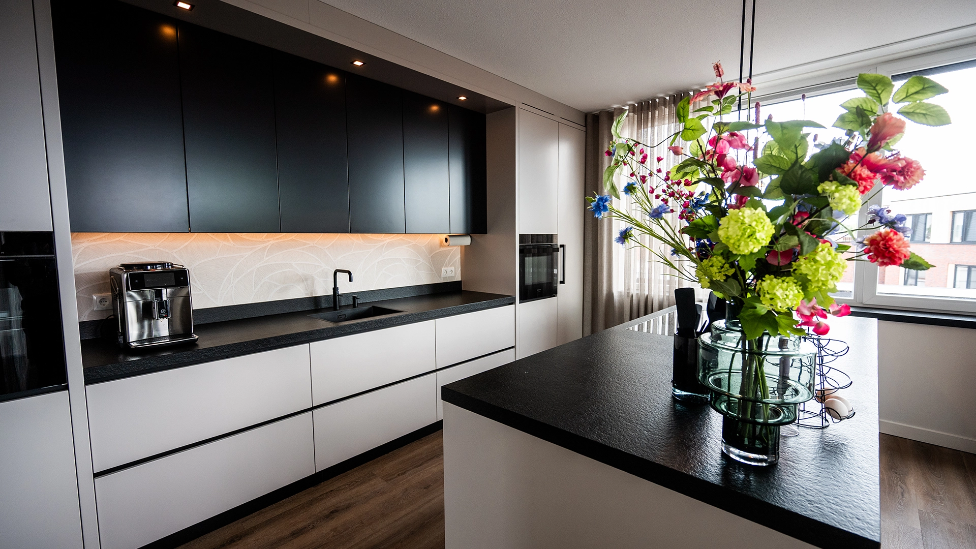 Strakke moderne keuken met zwarte en witte kasten, donker keukenblad, ingebouwde apparatuur en kookeiland met bloemenboeket, ontworpen door Nouk Interiors.