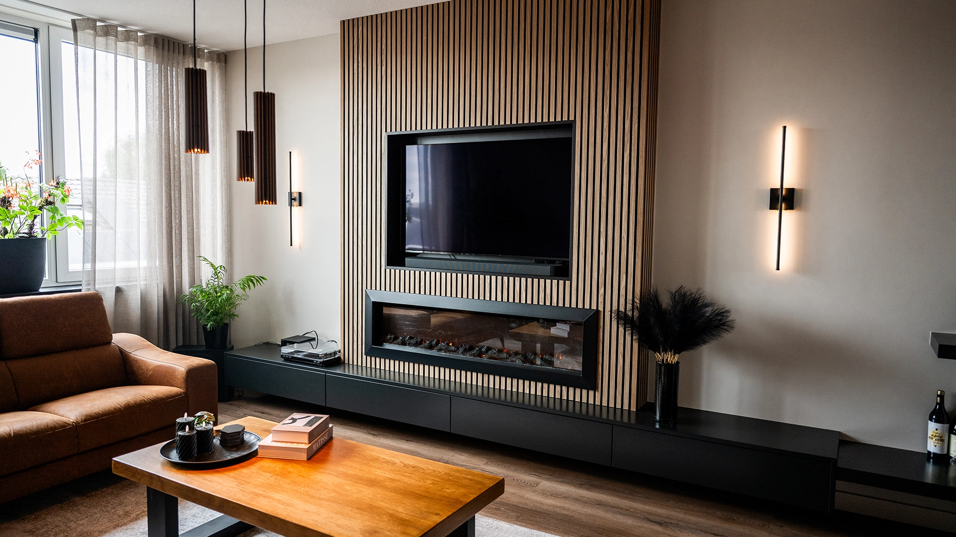 Moderne woonkamer met houten lattenwand, ingebouwde haard en tv, leren bank, houten salontafel en stijlvolle verlichting, ontworpen door Nouk Interiors.
