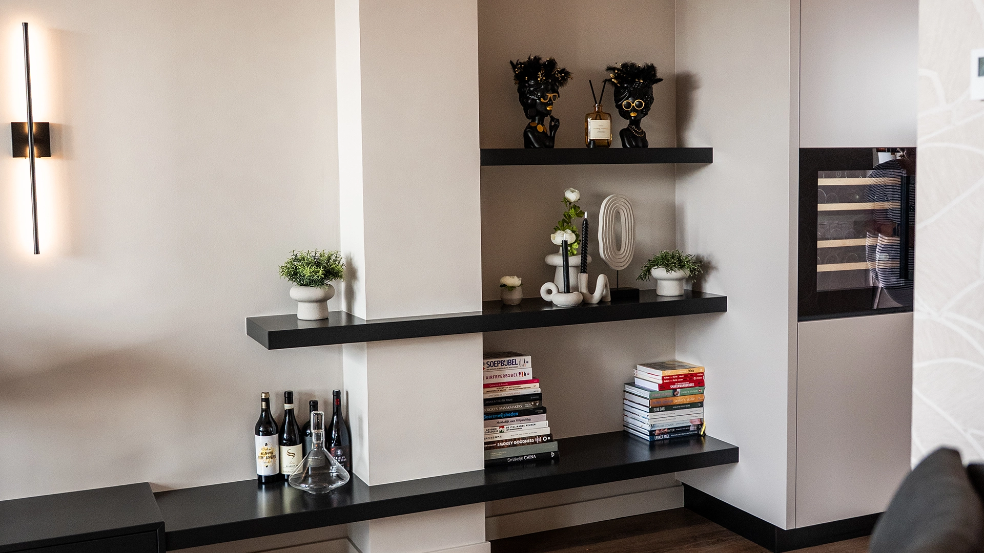 Stijlvolle woonkamer met zwarte wandplanken, decoratieve accessoires, boeken en wijnflessen, onderdeel van modern interieurontwerp door Nouk Interiors.
