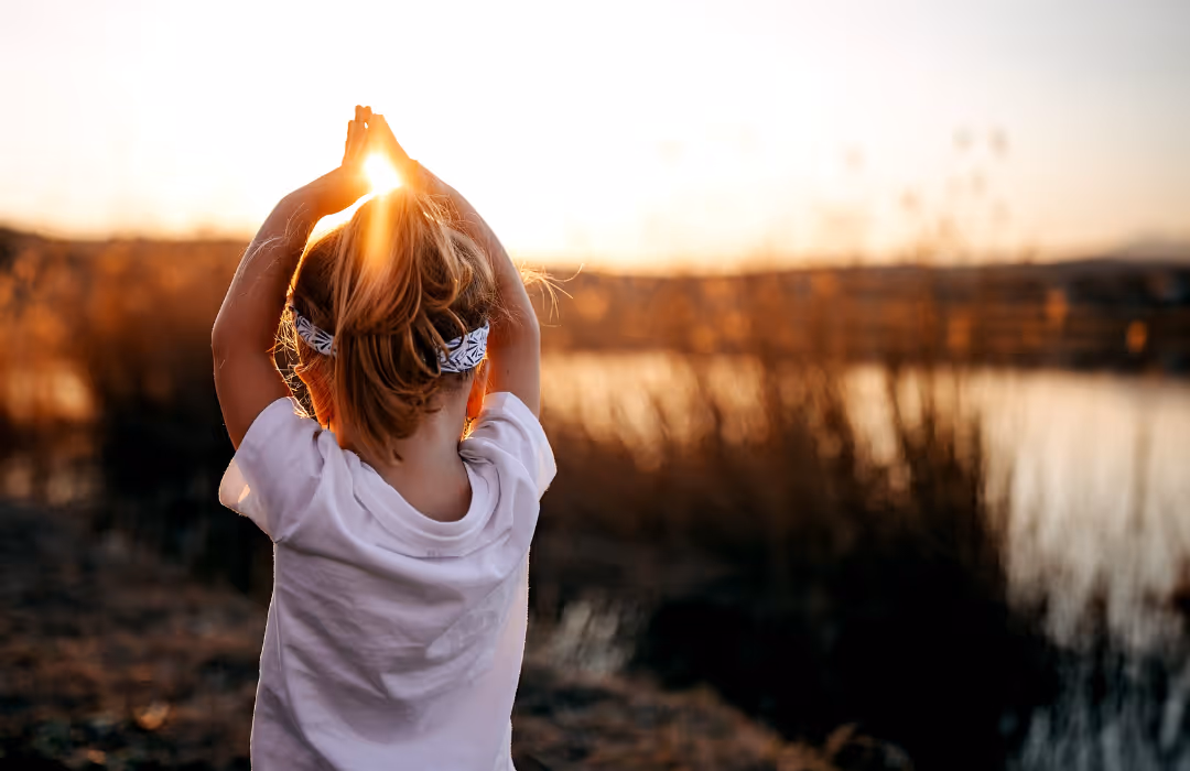 Kind führt Yogaübung draußen bei Sonnenuntergang am Wasser durch.