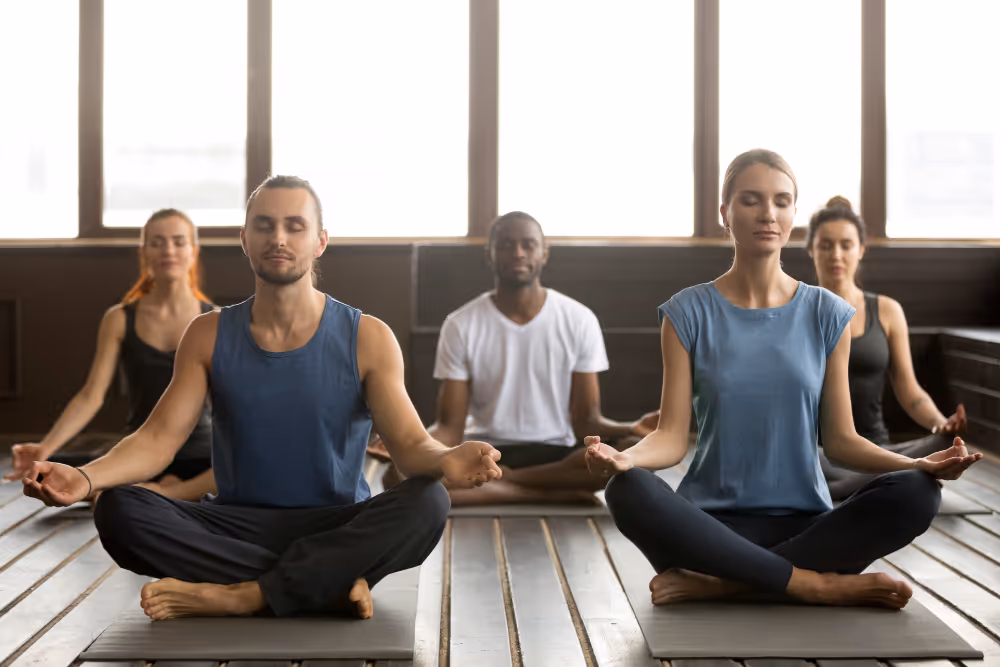 Gruppe von fünf Personen sitzt im Schneidersitz mit geschlossenen Augen und meditierendem Ausdruck in einem hellen Raum mit großen Fenstern.