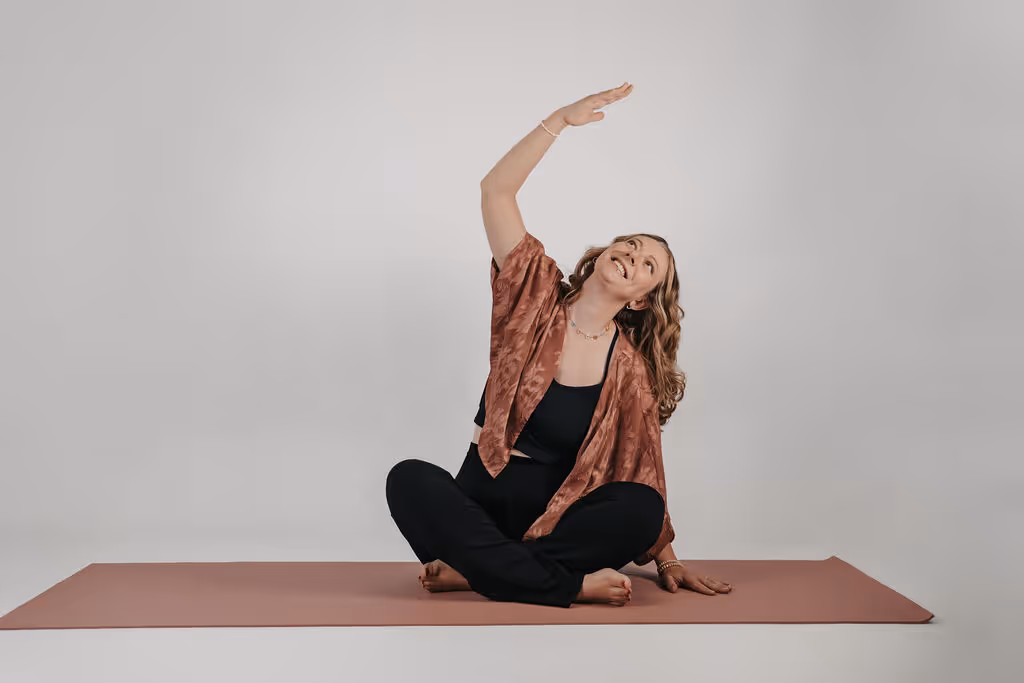 Frau sitzt auf einer Yogamatte im Schneidersitz und streckt ihren rechten Arm nach oben, Blick nach oben gerichtet.