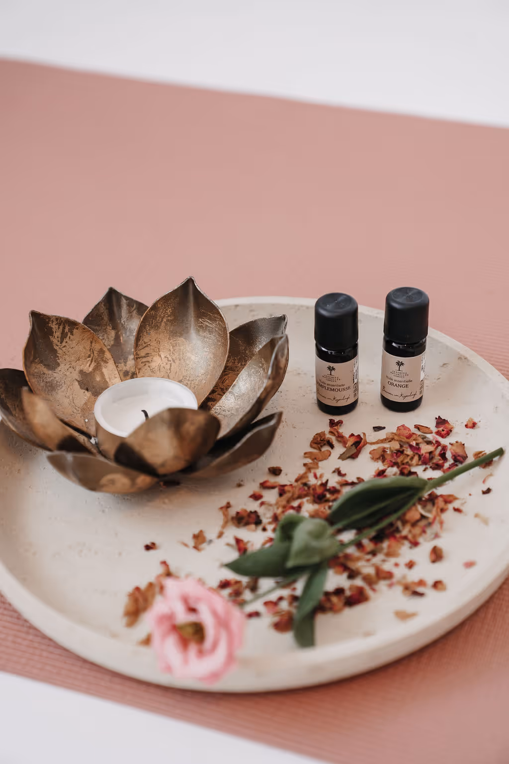 Runde Platte mit goldenem Kerzenhalter, zwei Aromaflaschen, getrockneten Blütenblättern und einer rosa Blume auf rosa Unterlage.