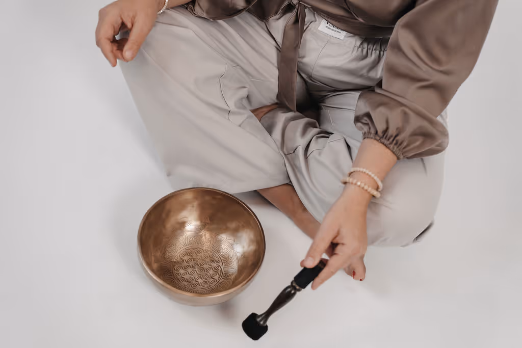Person sitzt im Schneidersitz auf weißem Boden und hält Schlegel neben bronzefarbener Klangschale mit eingraviertem Muster.