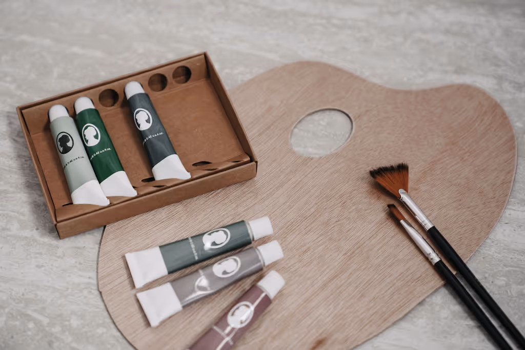 Holzmalpalette mit sechs Farbtuben und drei Pinsel auf grauem Untergrund.