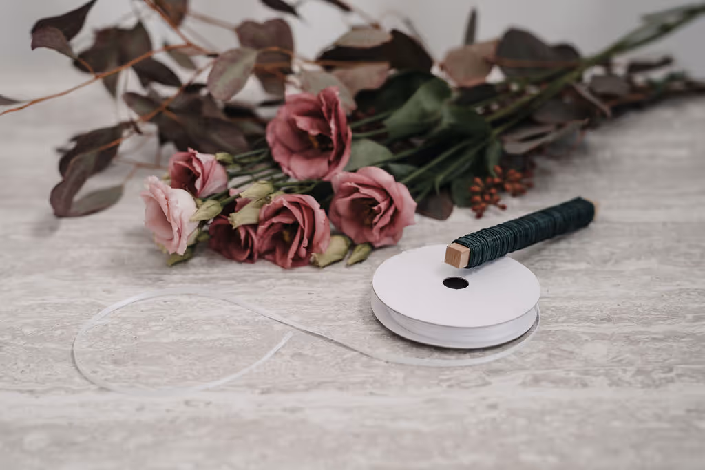 Strauß mit rosa und lila Blüten, daneben weiße Blumendrahtrolle und grüner Blumendraht auf Holztisch.