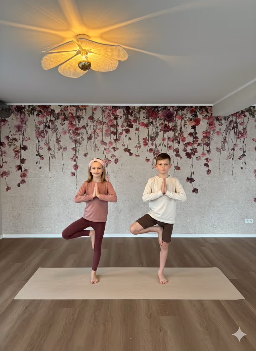 Ein Mädchen und ein Junge stehen auf einer Yogamatte in der Baum-Pose mit zusammengelegten Händen in einem Raum mit floraler Wanddekoration und Deckenleuchte.