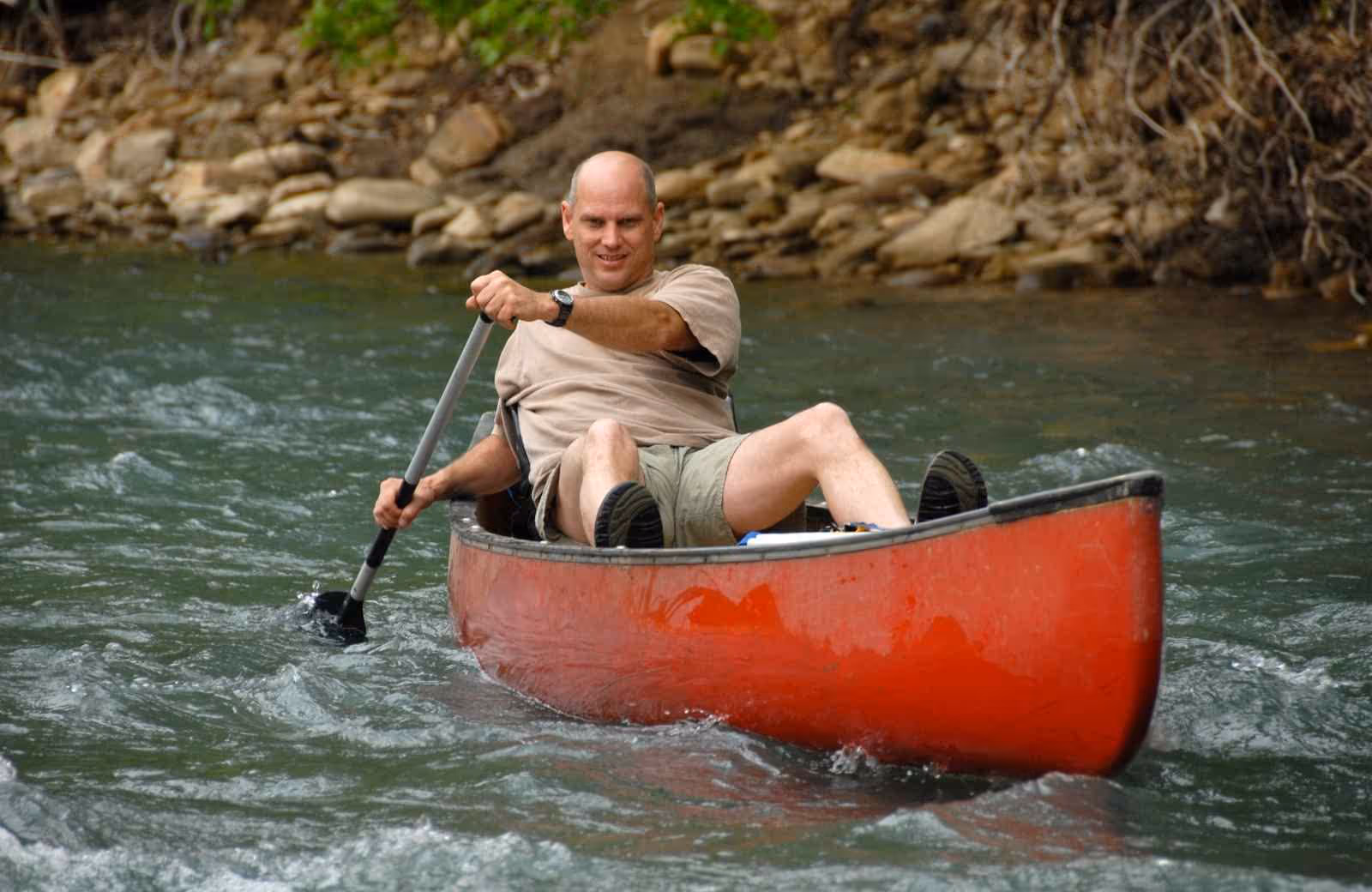 Homme pagayant seul dans un canoë rouge sur une rivière aux eaux vives, entourée de rochers et de végétation