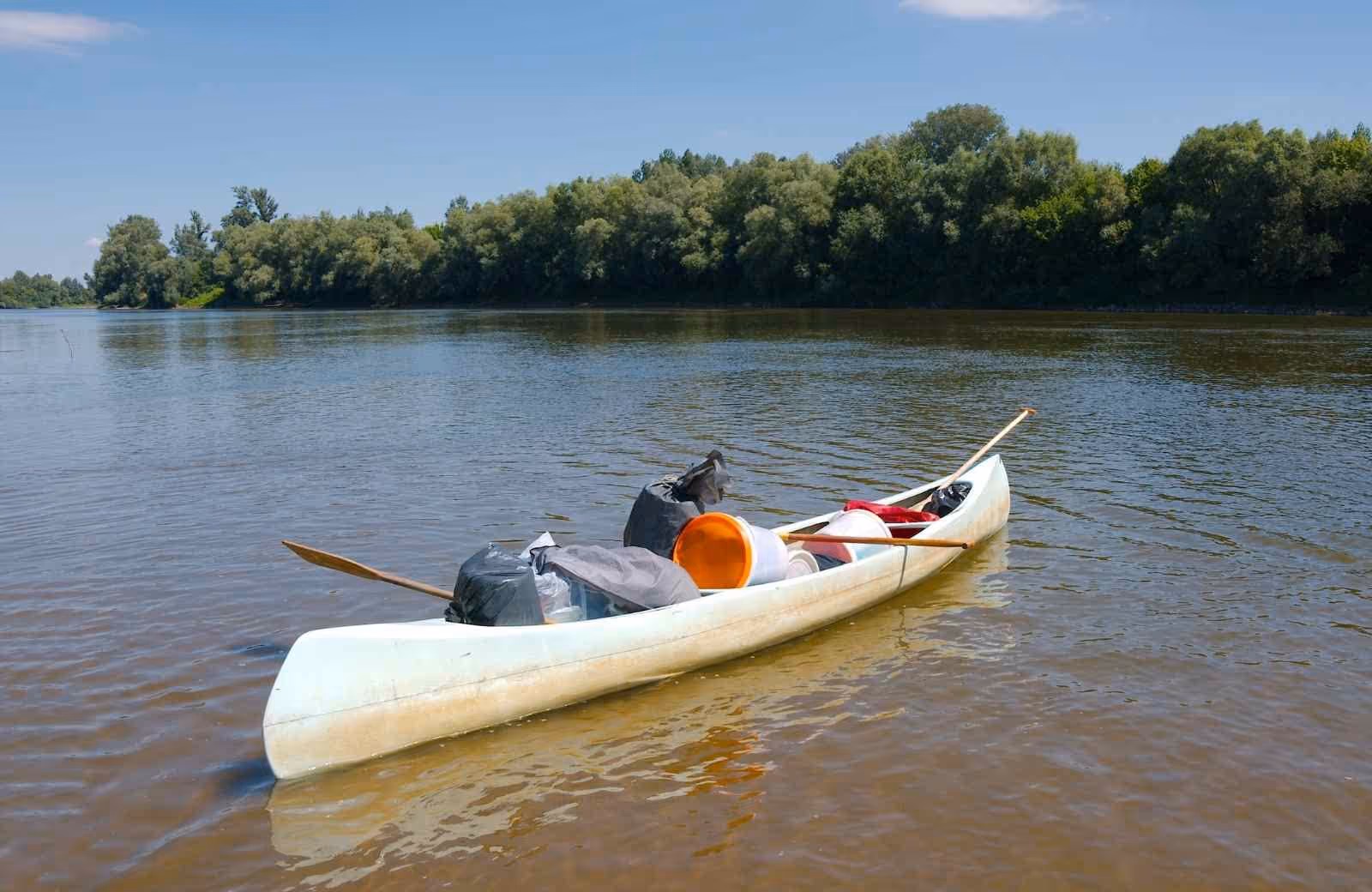 Canoë blanc chargé de matériel et de bidons étanches, flottant sur une rivière calme entourée d’arbres.