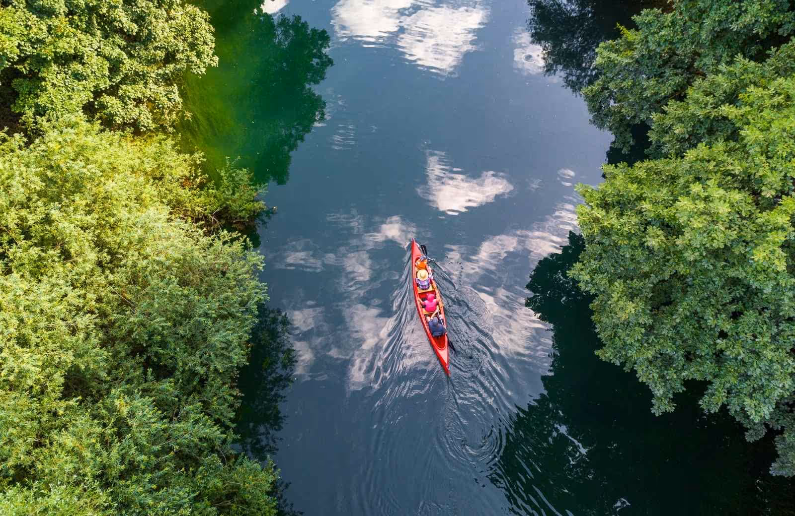 Vue aérienne d’un canoë rouge avec deux personnes pagayant sur la Loire, entourée d’une végétation dense et reflet du ciel sur l’eau calme.