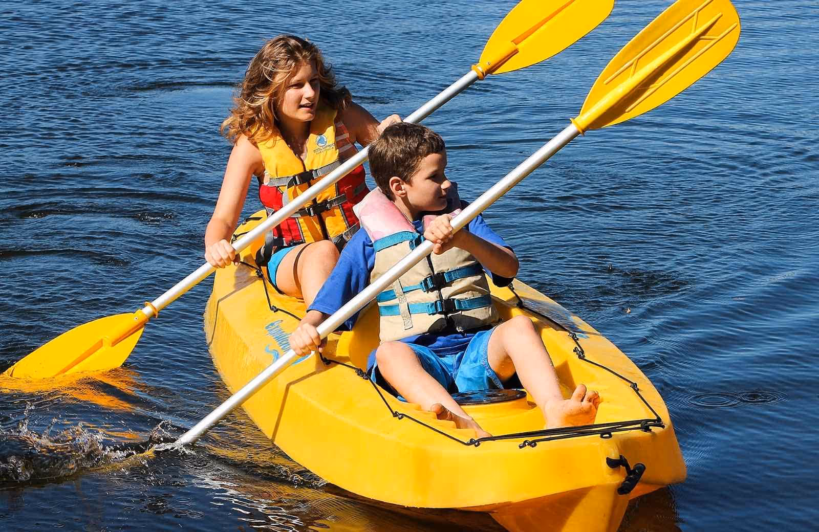 Femme et enfant pagayant ensemble dans un kayak jaune sur une eau calme, tous deux équipés de gilets de sauvetage.