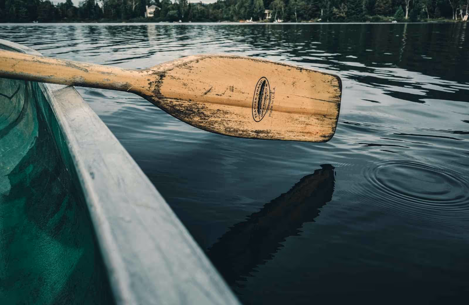 Vue rapprochée d’une pagaie en bois posée sur le bord d’un canoë vert flottant sur un lac calme.