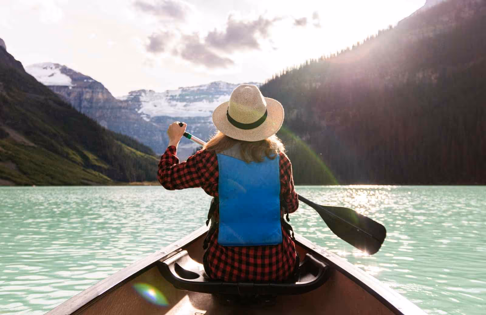 Femme en gilet de sauvetage et chapeau pagayant dans un canoë sur un lac turquoise entouré de montagnes et de forêts.