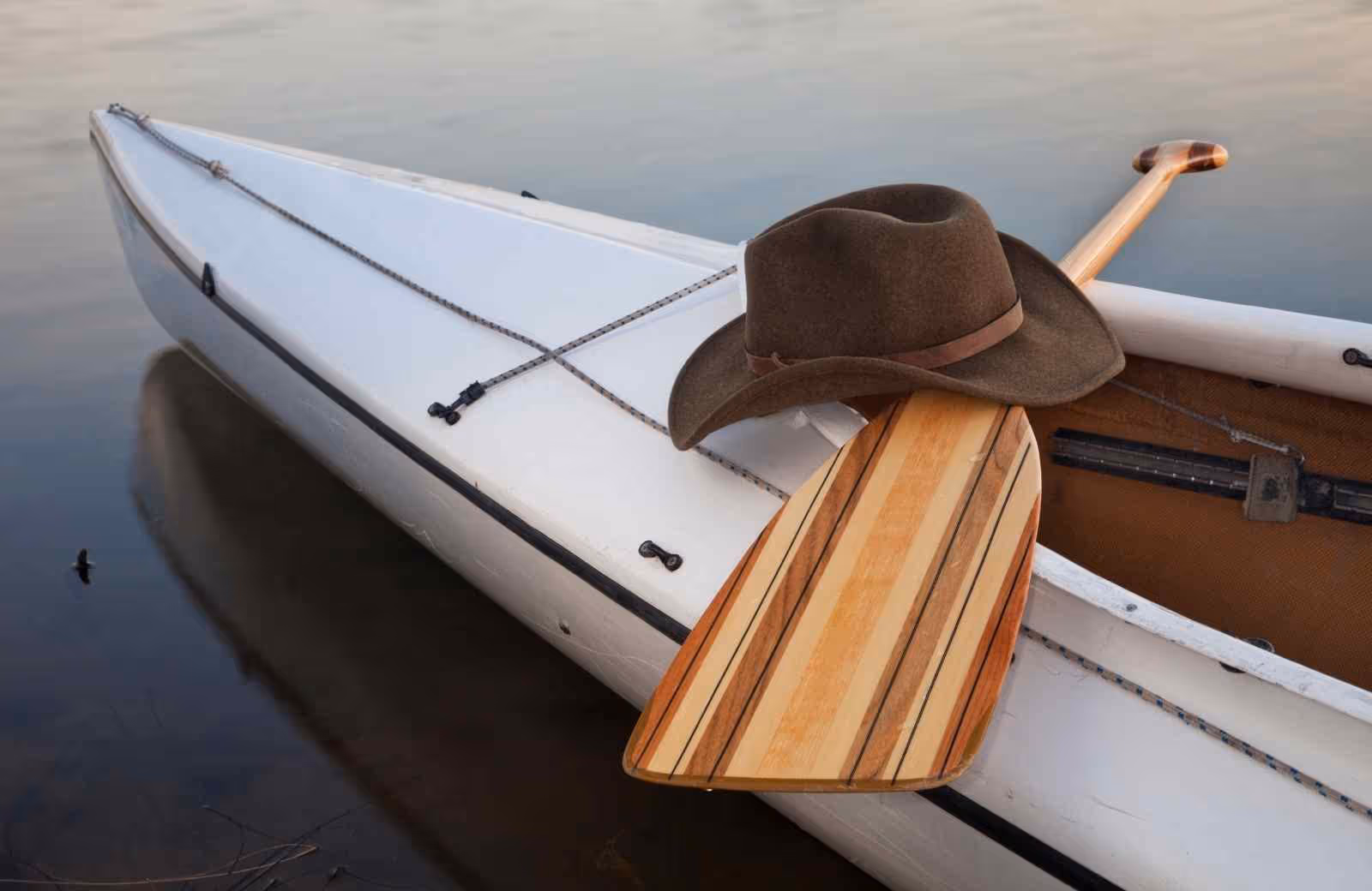 Canoë blanc au bord de l’eau avec une pagaie en bois rayé et un chapeau posé dessus, en fin de journée calme.