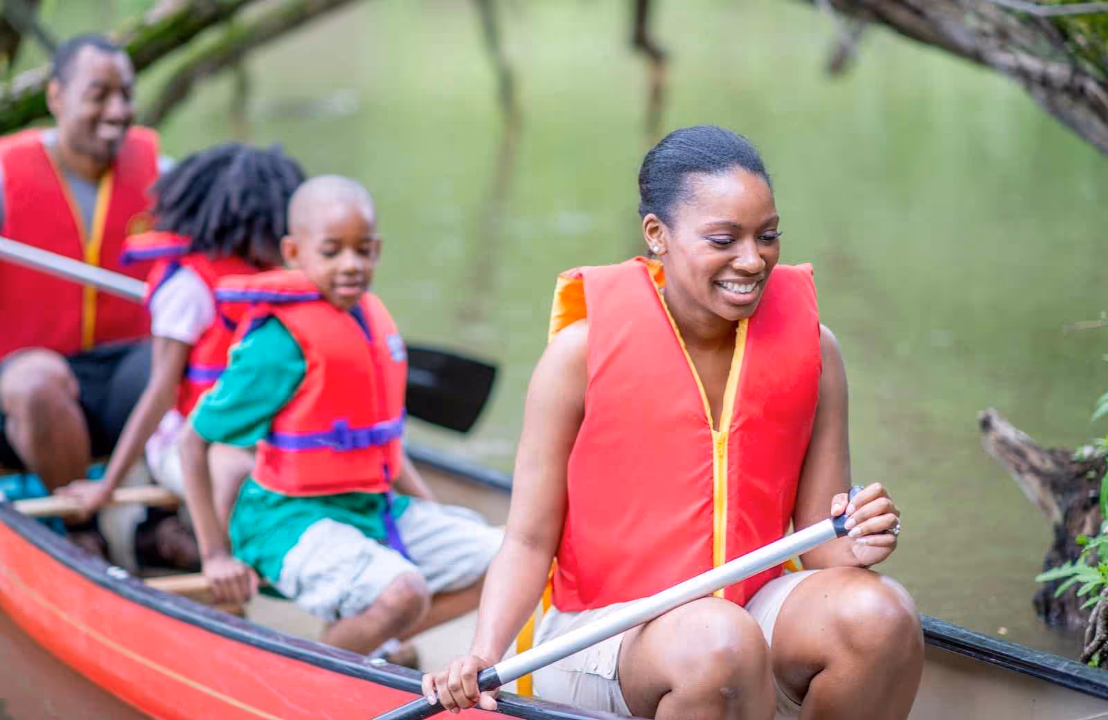 Famille souriante pagayant dans un canoë sur une rivière calme, tous équipés de gilets de sauvetage rouges.