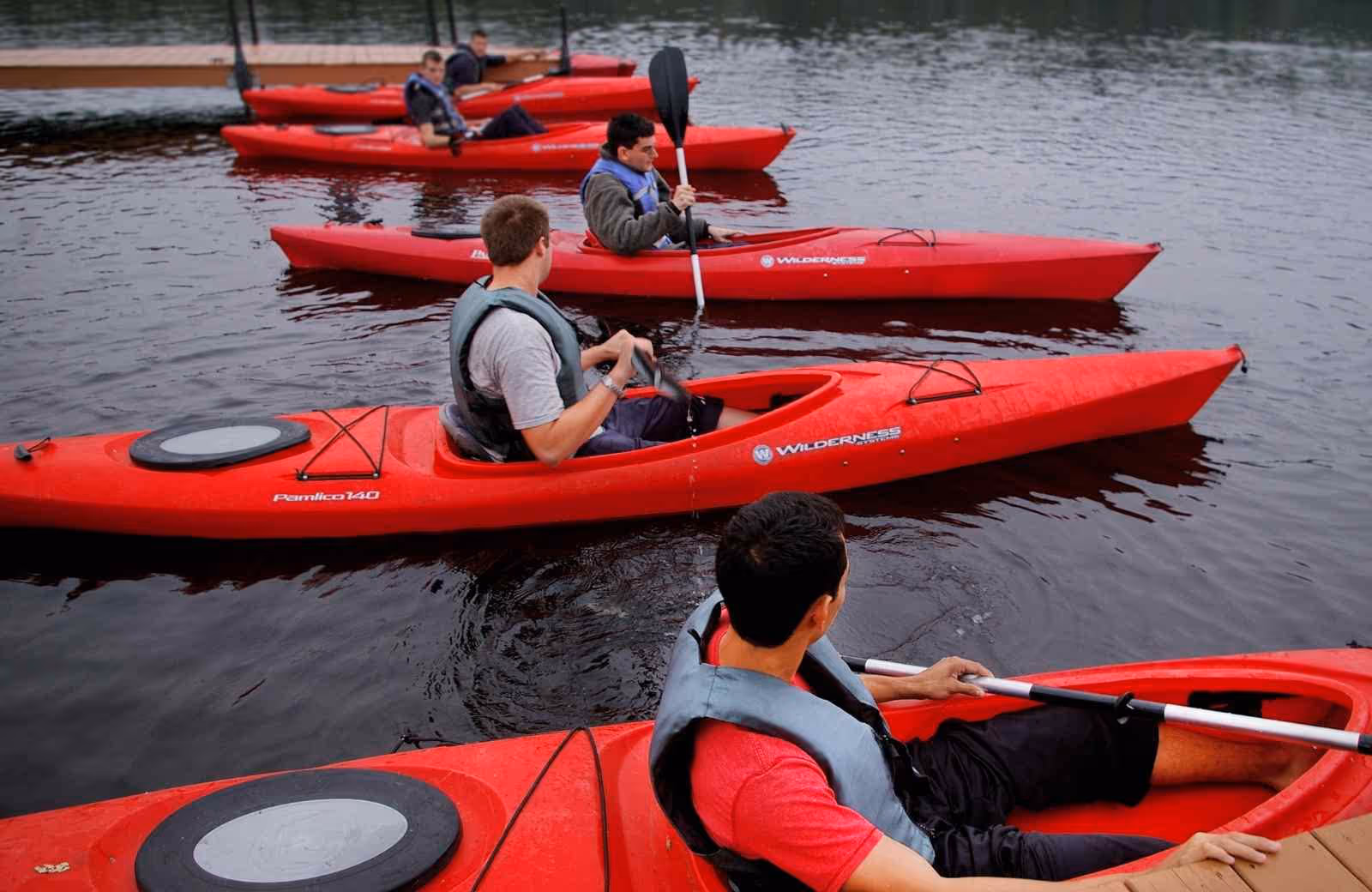 Groupe d’hommes en gilets de sauvetage s’entraînant au maniement du kayak rouge sur un plan d’eau calme, près d’un ponton.