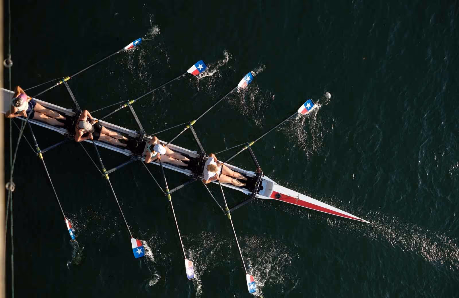 Vue aérienne d’une équipe d’aviron en pleine synchronisation, pagayant sur une eau sombre avec des rames ornées de drapeaux.