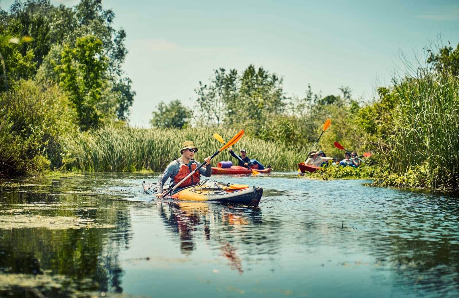 Groupe de kayakistes pagayant sur une rivière entourée de végétation dense par une journée ensoleillée, avec un pagayeur en tête portant un chapeau et des lunettes de soleil.