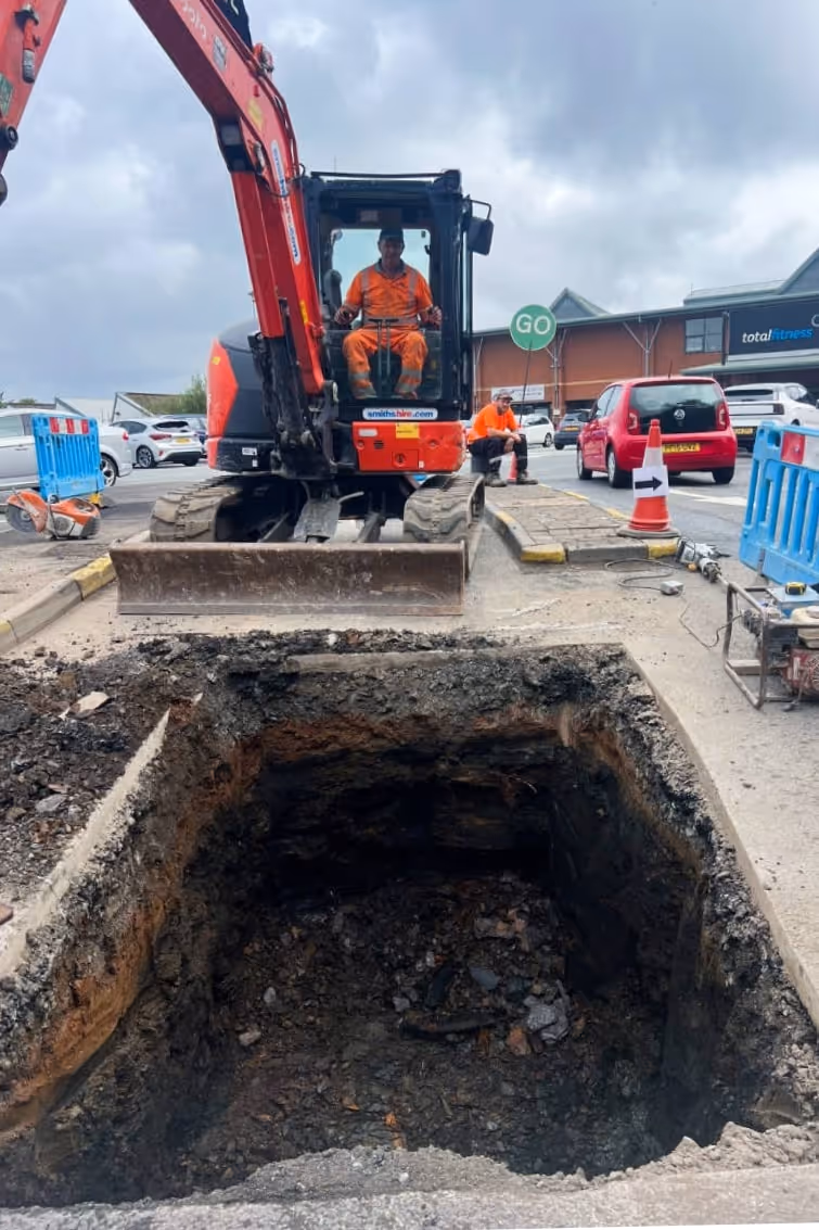 Excavator digging large hole in parking lot with construction worker