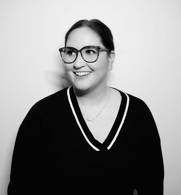 Black and white portrait of Meredith Murray smiling and looking to the side, wearing glasses and a dark V-neck sweater with a necklace.