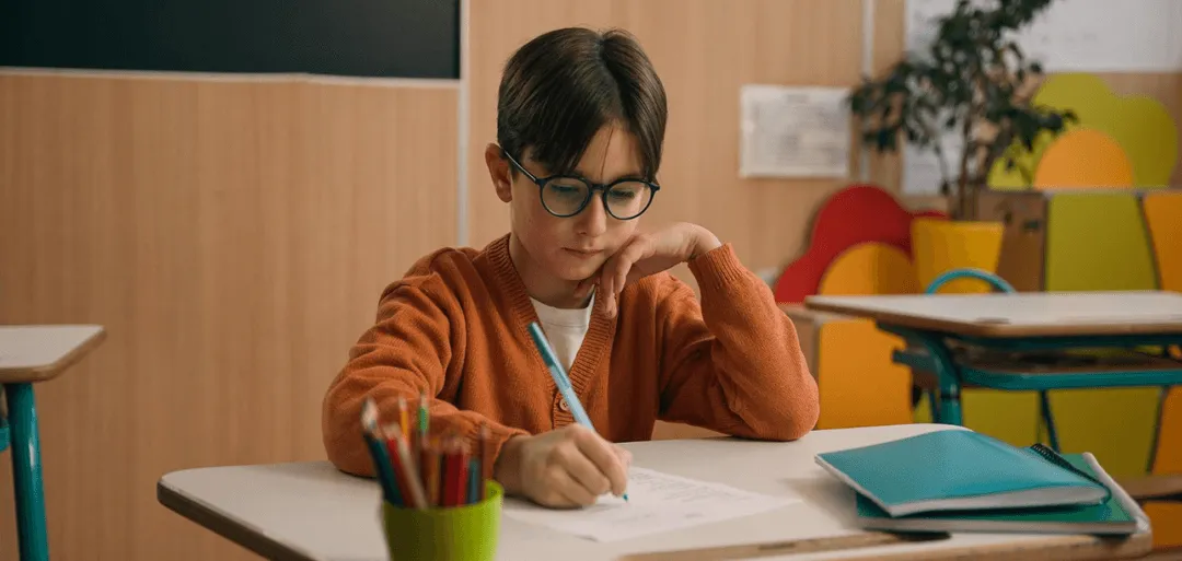 A young boy sitting at a desk with a book and pencils.