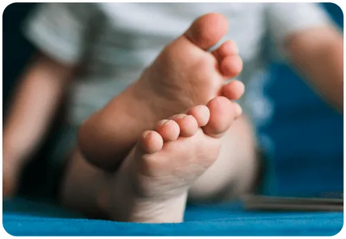 Close-up of a newborn baby's tiny feet with pink toes, lying on a soft blue blanket or surface, with blurred figures of adults visible in the background
