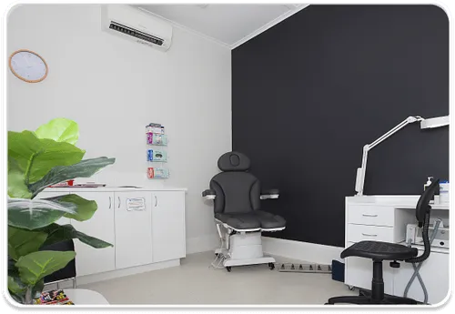 Modern dental office interior featuring a black dental chair with adjustable arm, white cabinets along the wall, dental equipment including an overhead light and instruments, a large leafy green plant in the corner, and educational posters on the white walls. The room has clean white flooring and a dark accent wall.