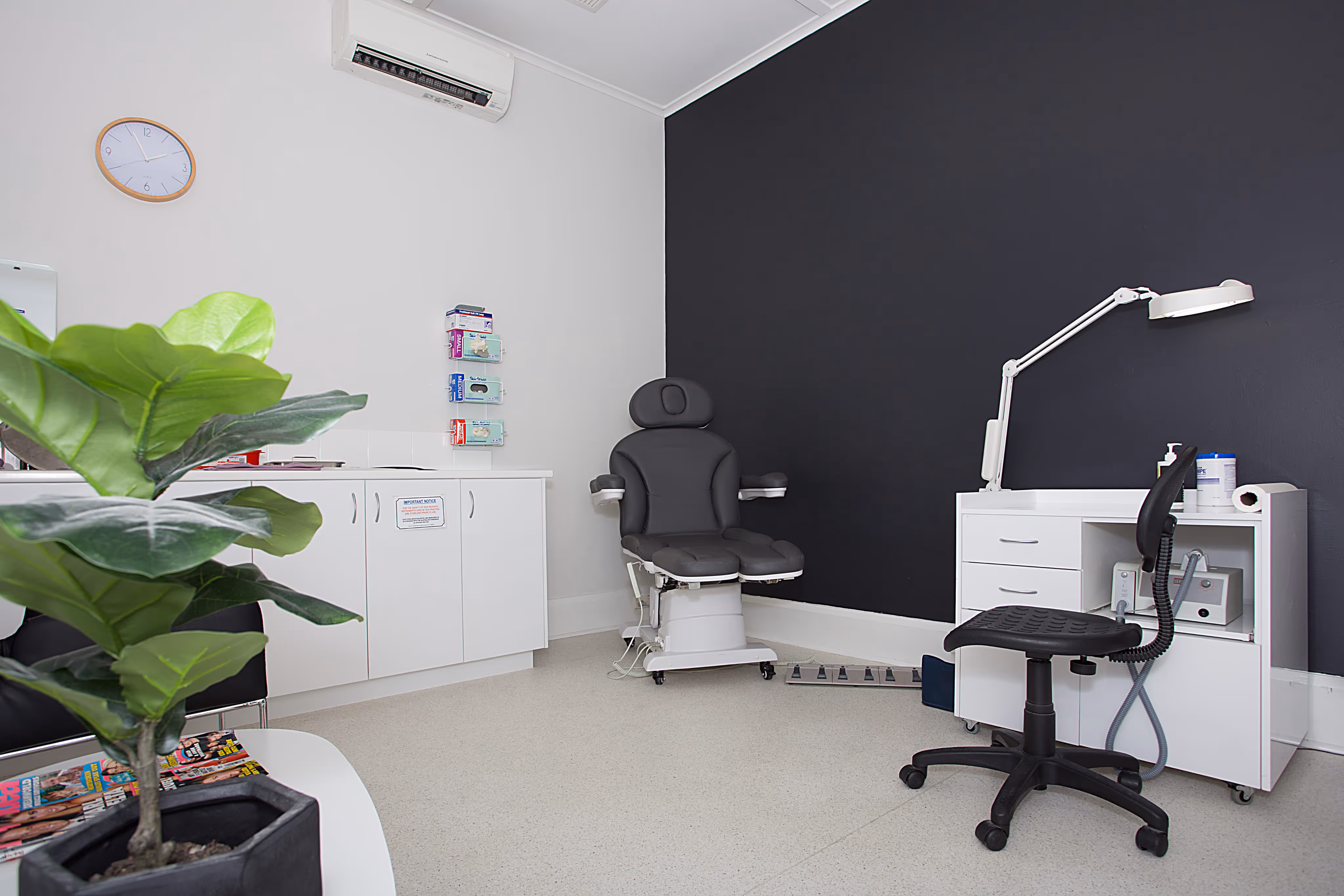 Modern medical examination room with a black treatment chair, white medical equipment cart, adjustable LED examination light, white storage cabinets, and a large green plant in the corner. The room features a dark accent wall, white walls with a round clock, and appears clean and professionally designed.
