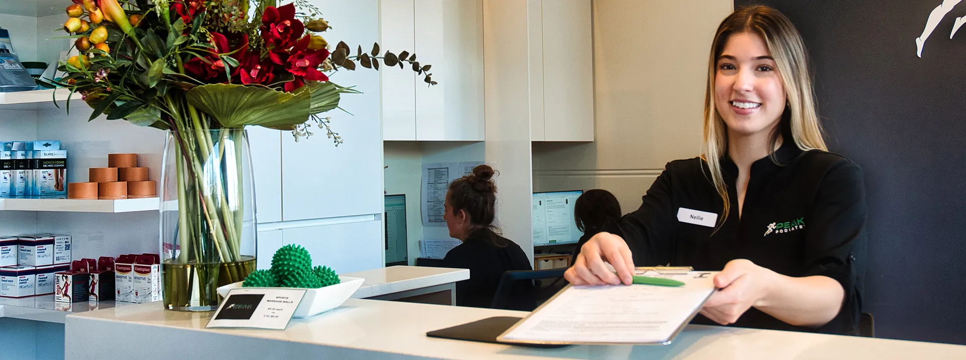 A friendly female receptionist smiling and extending a clipboard with forms to a patient.