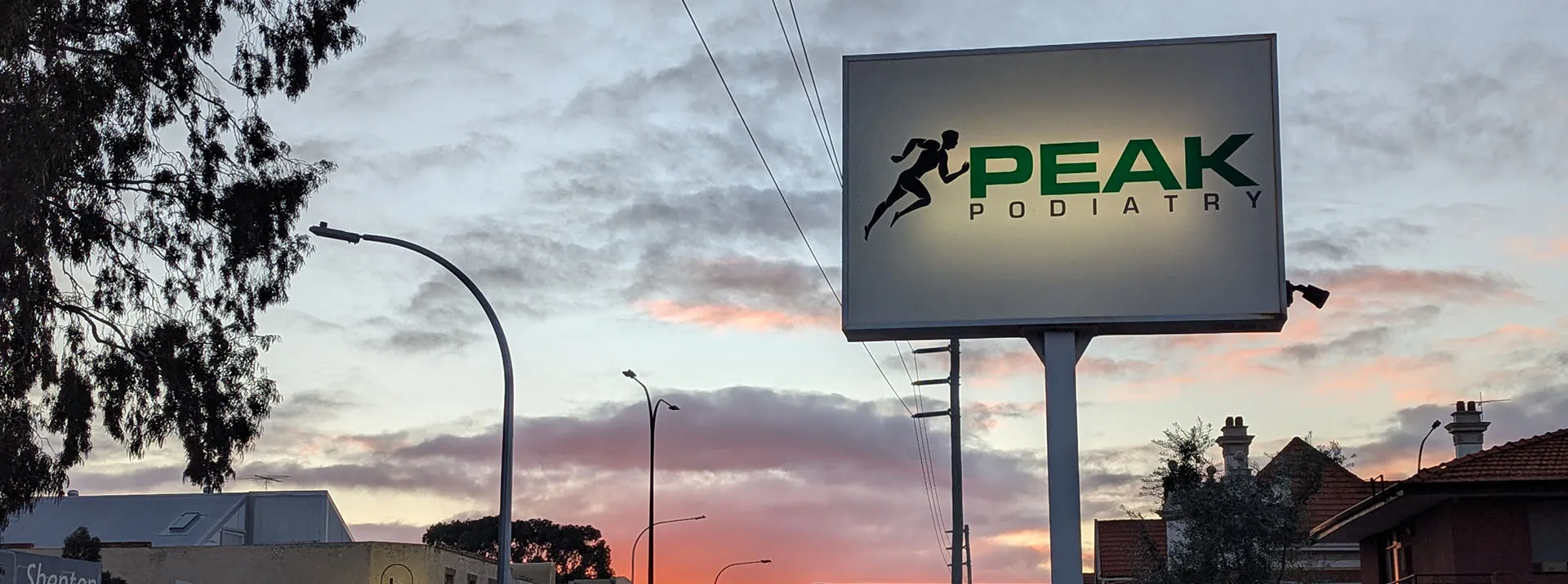 An illuminated billboard for Peak Podiatry with a runner logo against a colorful sunset sky.