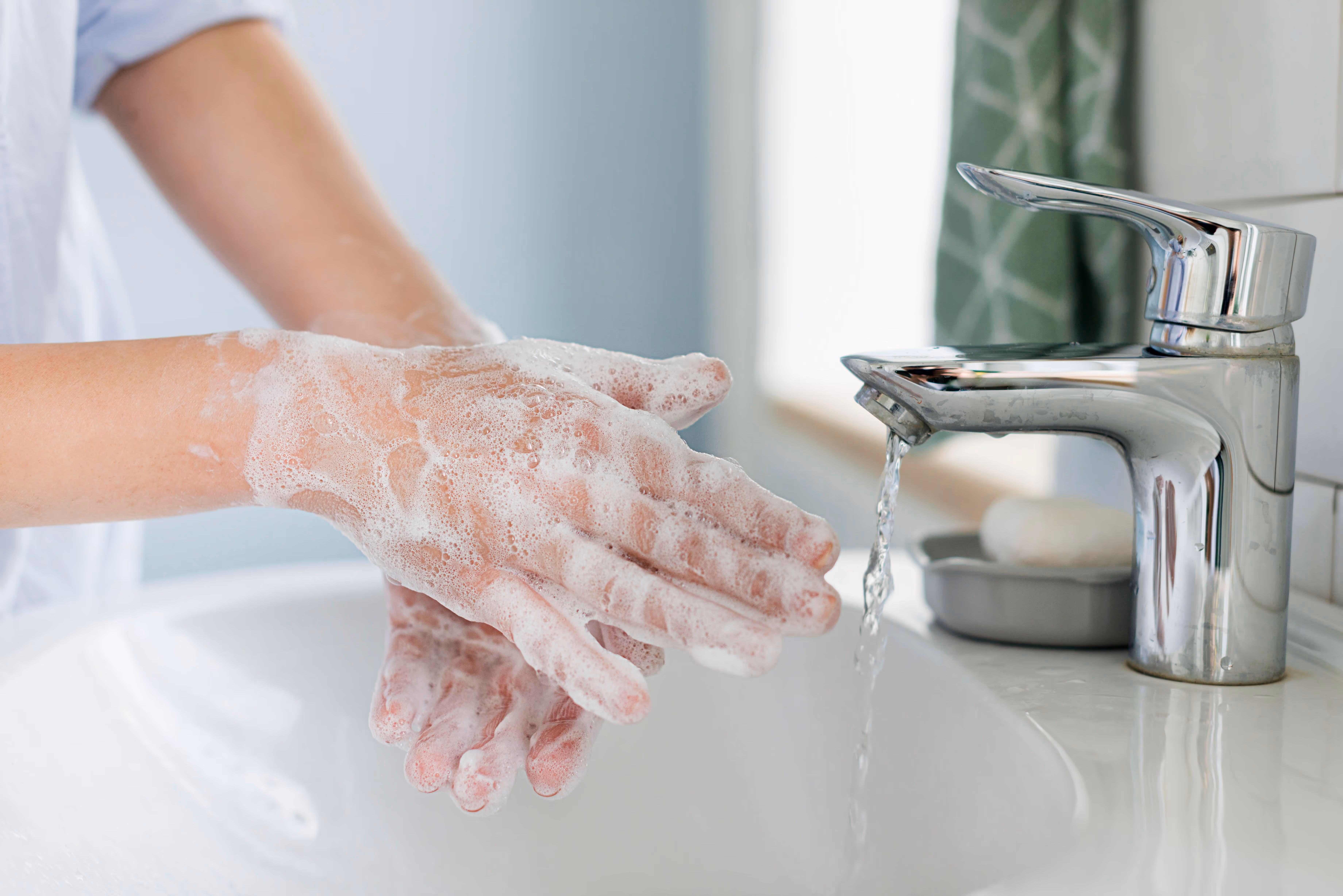 A person's hands covered in white soap suds are held under a stream of water flowing from a modern chrome faucet into a white sink
