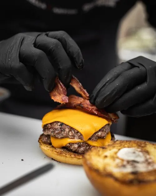 A person in black gloves is putting bacon on a cheeseburger.