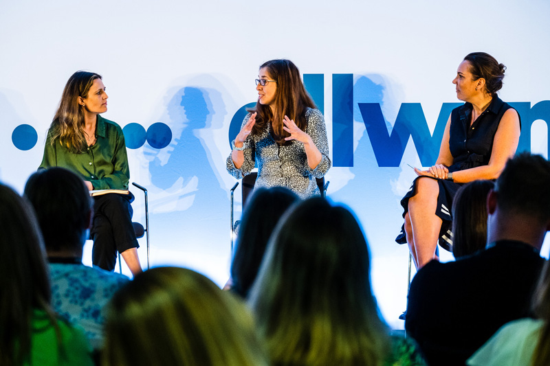 Two women and a host participate in a discussion during a panel session at a conference. One of the women is speaking while the others are listening attentively.