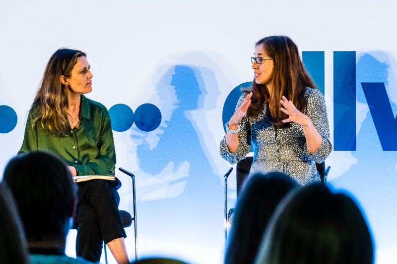 Two women are engaged in a conversation on stage at an event, with one gesturing while speaking. The background features abstract, colorful shapes.