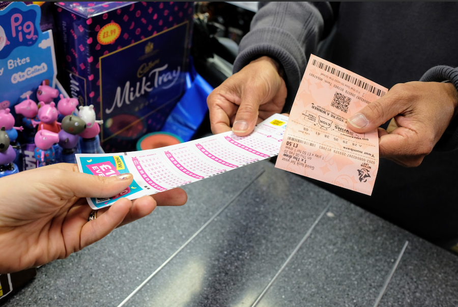 A person hands a lottery ticket to another person at a store checkout. The person receiving the ticket holds a completed lottery slip, while a box of Cadbury Milk Tray is visible in the background.