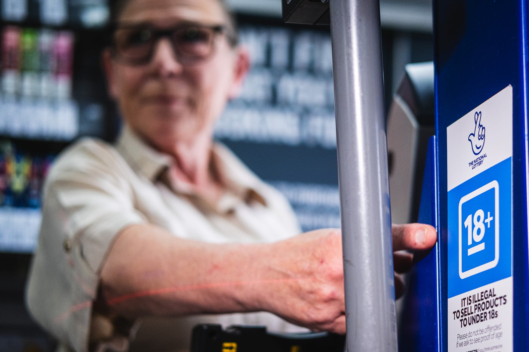 A person holds a National Lottery kiosk with an age verification label on it. The label highlights that lottery products are for individuals aged 18 and over.