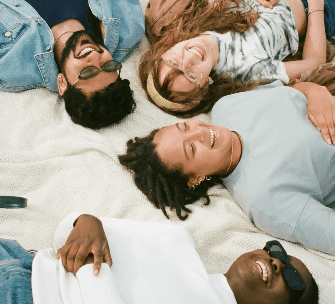 A group of four friends lying on a blanket outdoors in a circle with their heads close together, all smiling and laughing. They are casually dressed, wearing sunglasses, and appear to be enjoying a relaxed, joyful moment.