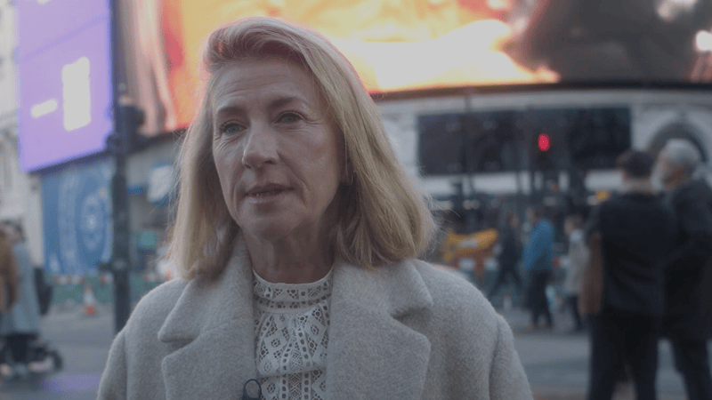 A woman standing outside in a busy city environment, likely in London, with a large digital screen in the background. She has blonde hair, is wearing a light-colored coat, and looks directly at the camera with a neutral expression. People are walking in the background, and the scene captures the hustle and bustle of the city.