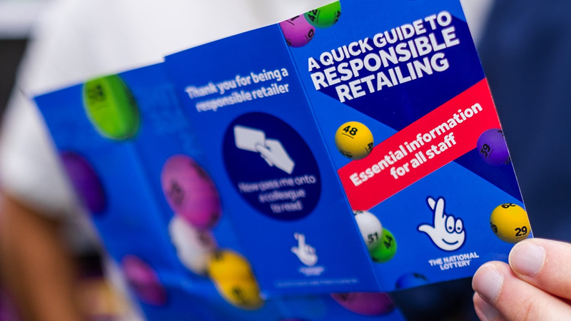 A close-up of a person holding a leaflet titled "A Quick Guide to Responsible Retailing," with colorful lottery balls in the background. The leaflet provides essential information for lottery retailers.