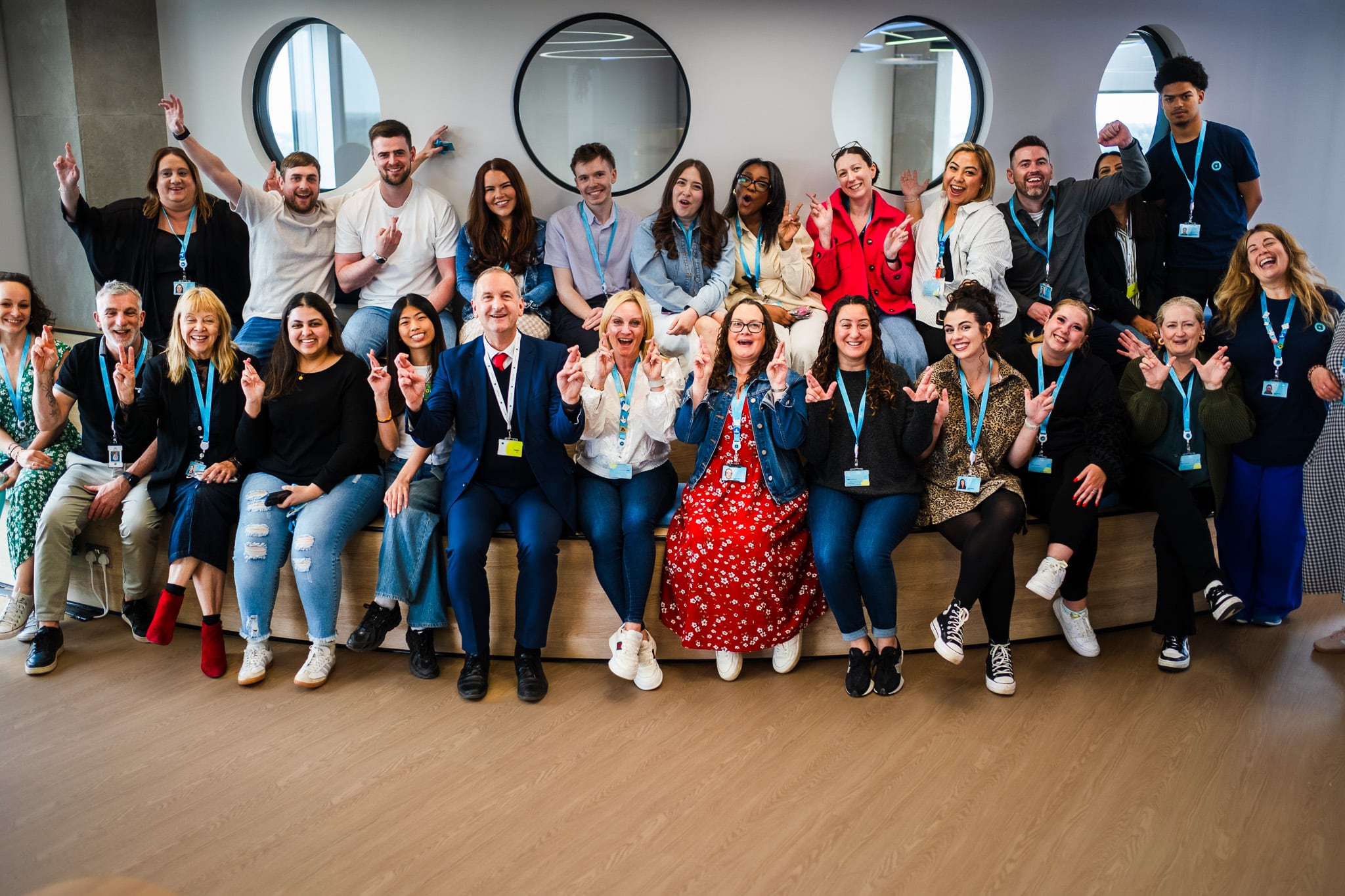 A group photo of a diverse team of employees, standing and sitting in a modern office environment. Everyone is smiling and making the 'I love you' gesture with their hands. The team is wearing lanyards and badges, and the space features circular windows in the background