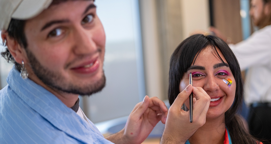 This image shows a person applying makeup on another person’s face. The person getting the makeup done has bright, colorful makeup on their eyes and a flag design on their cheek. The one applying the makeup is smiling and looking at the camera. There’s a sense of fun and creativity in this moment.