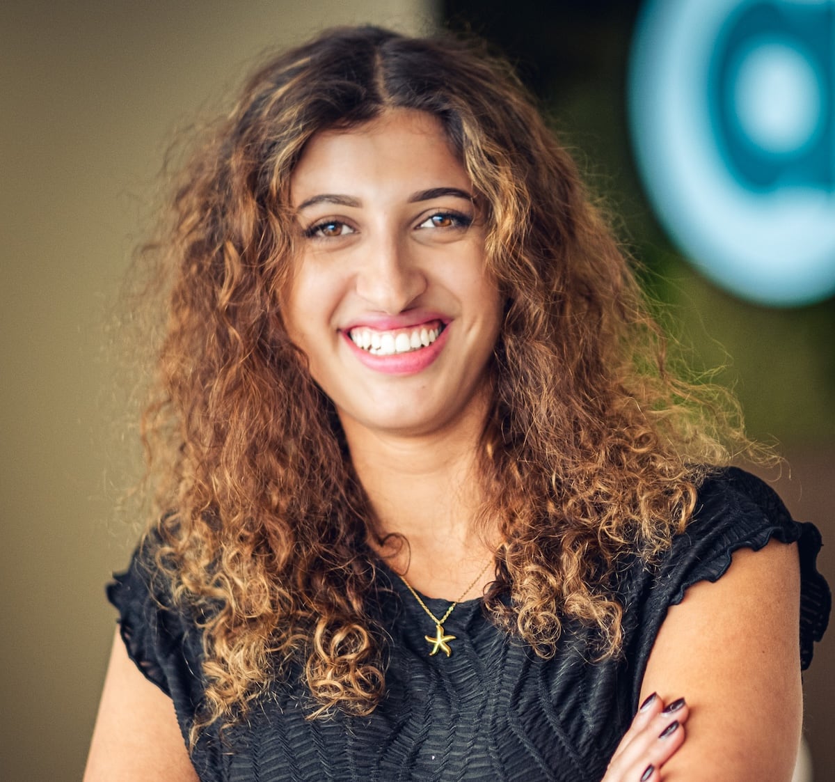 A woman named Sophia, smiling and standing indoors with her arms crossed. She has curly hair and is wearing a black top. She is wearing a gold starfish necklace and has a friendly expression on her face.