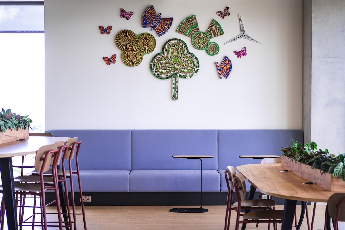 an interior space, likely a modern office or lounge area. The wall is decorated with colorful, abstract artwork featuring circular patterns and butterflies. The furniture consists of a sleek, modern blue couch with a minimalist table beside it, as well as a long wooden table with plants along its edges. The decor gives the space a contemporary and artistic feel