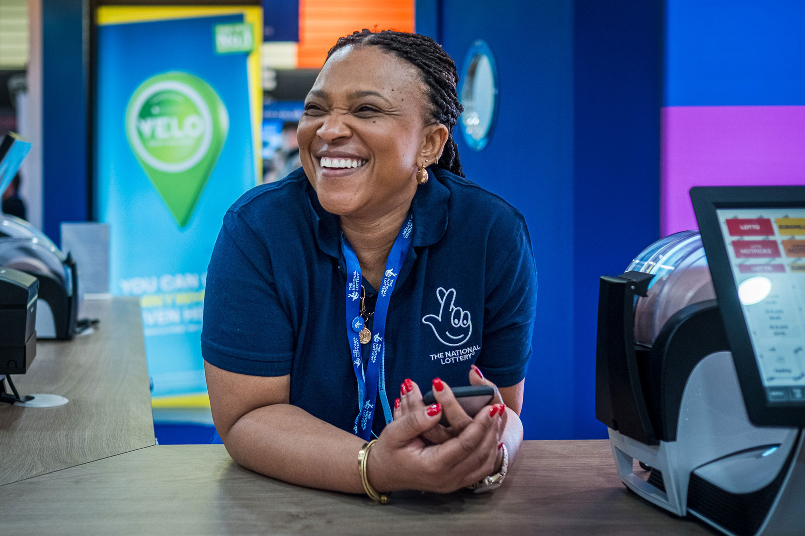 a modern office reception area with a welcoming vibe. The background features a vibrant green moss wall, and the company logo, "Allwyn," is prominently displayed. The digital screen above the reception desk displays the message: "Welcome to Clarendon Works, Allwyn UK." The reception desk is sleek, and there are people interacting and working at the desk, with a few standing near the entrance. The space has a clean and contemporary design.