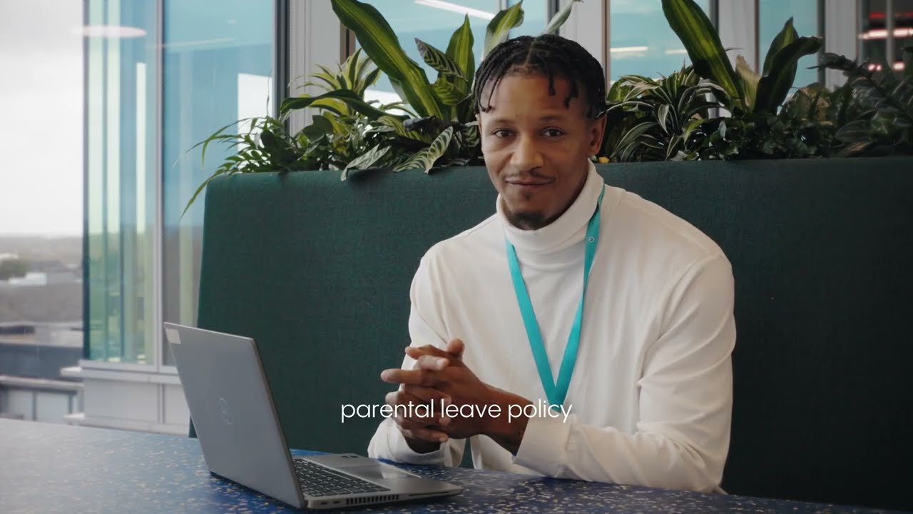A woman standing outside in a busy city environment, likely in London, with a large digital screen in the background. She has blonde hair, is wearing a light-colored coat, and looks directly at the camera with a neutral expression. People are walking in the background, and the scene captures the hustle and bustle of the city.