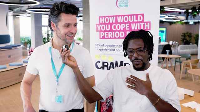 a modern office reception area with a welcoming vibe. The background features a vibrant green moss wall, and the company logo, "Allwyn," is prominently displayed. The digital screen above the reception desk displays the message: "Welcome to Clarendon Works, Allwyn UK." The reception desk is sleek, and there are people interacting and working at the desk, with a few standing near the entrance. The space has a clean and contemporary design.