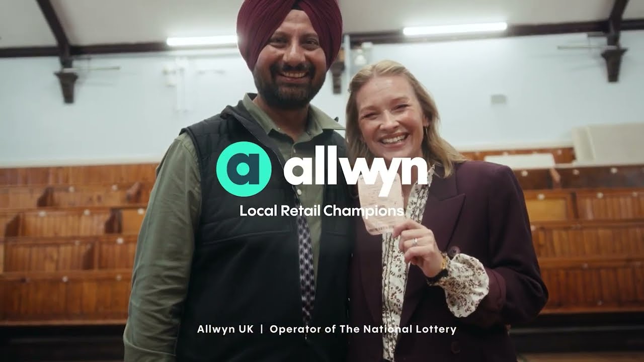 a modern office reception area with a welcoming vibe. The background features a vibrant green moss wall, and the company logo, "Allwyn," is prominently displayed. The digital screen above the reception desk displays the message: "Welcome to Clarendon Works, Allwyn UK." The reception desk is sleek, and there are people interacting and working at the desk, with a few standing near the entrance. The space has a clean and contemporary design.
